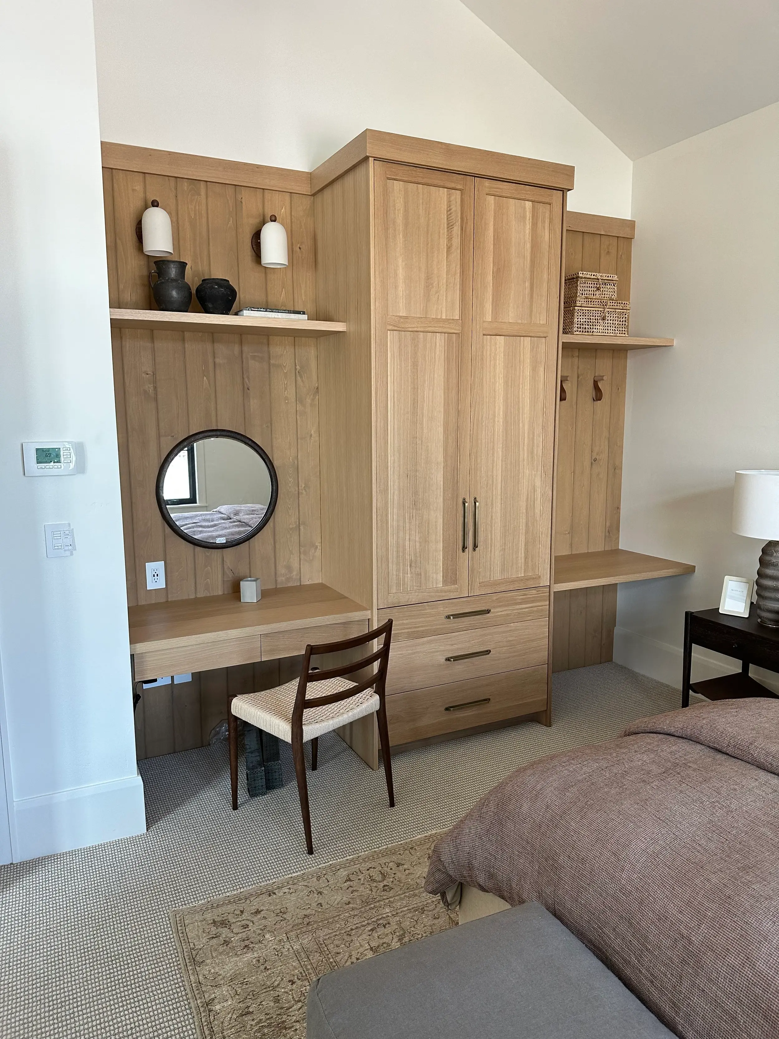 Light oak bedroom wardrobe with paneled doors and drawers beside a built-in desk nook with round mirror and tongue-and-groove paneling.
