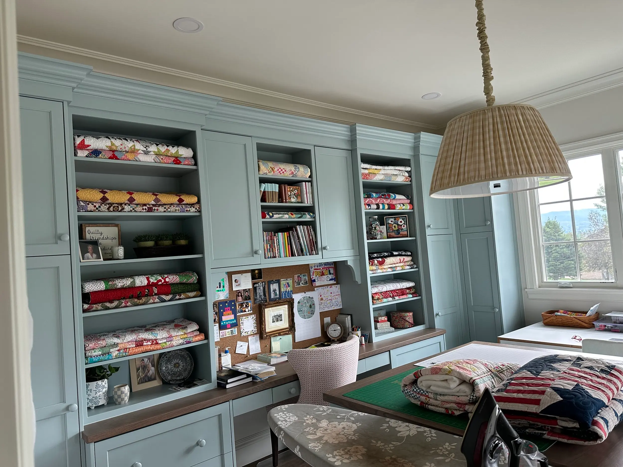 Custom craft room built-ins with open quilt storage shelving, integrated desk, and cabinetry finished in a soft blue-gray tone.